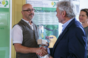 Sebastian Körnig von Public2Science nimmt den Umweltpreis 2022 von Dr. Willy Boss, dem Vorstandsvorsitzenden der SUNK, entgegen. Daneben wartet Dr. Nele Herkt, SUNK-Geschäftsführerin, darauf zu gratulieren.